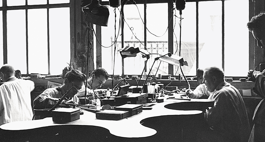 . Michael Darde, left foreground, with his father, Emile, at right, photographed in 1960 in the workshop at 12 rue Saint-Gilles in the Marais. Darde put in large conservatory windows so the workers could see by natural light. The workshop resembled a laboratory where craftsmen labored in white smocks, each secluded in his honeycomb cell. The senses were assaulted on every front: heat from the welding stations, light from the windows, the pervasive odor of cigarettes and the burnt stench of metal and resin. Among those who formed the precious jewels were Emile Darde, Maurice Groëné, Michel Darde, Gaëtan Bergonzoli, Pierre Vitasse, “old man Baillet,” Louis Berson, Jacques Ferré, Charles l’Hospital, Désiré l’Hospital and Roger. Credit: Darde Family Archives.