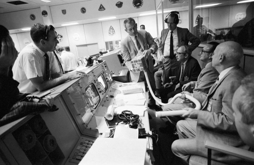 “Working the problem:”  Deke Slayton (checked jacket) shows the adapter devised to remove excess carbon dioxide from the Apollo 13 Lunar Module cabin, which was dramatized in the movie “Apollo 13, based on “Lost Moon” by Jim Lovell and Jeffrey Kluger. From left, members of Slayton's audience are flight director Milton L. Windler; deputy director/flight operations Howard W. Tindall; director/flight operations Sigurd A Sjoberg; deputy director/Manned Spaceflight Center Christopher C. Kraft; and director/Manned Spaceflight Center Robert R. Gilruth. Photograph courtesy of NASA.