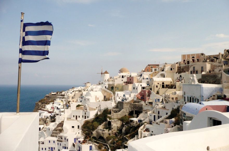 The Greek flag flies above the iconic, whitewashed buildings of Santorini.