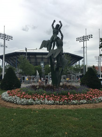 A sculpture of two dancers soars with Arthur Ashe Stadium in the background.