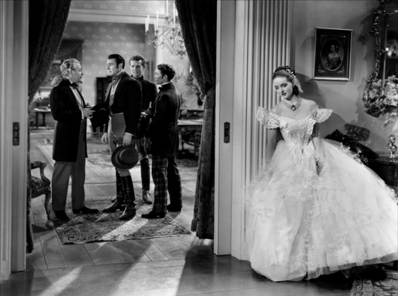 A vixenish Southern belle (Bette Davis) eavesdrops on her many admirers, who include George Brent (center) in “Jezebel” (1938). Film still used on the April 1938 cover of the National Board of Review Magazine.